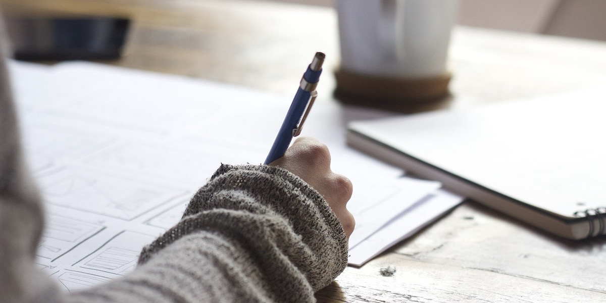 Designer's hand sketching layout wireframes on paper beside a notebook and mug
