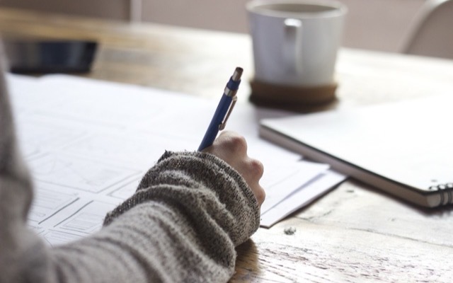 Designer's hand sketching layout wireframes on paper beside a notebook and mug